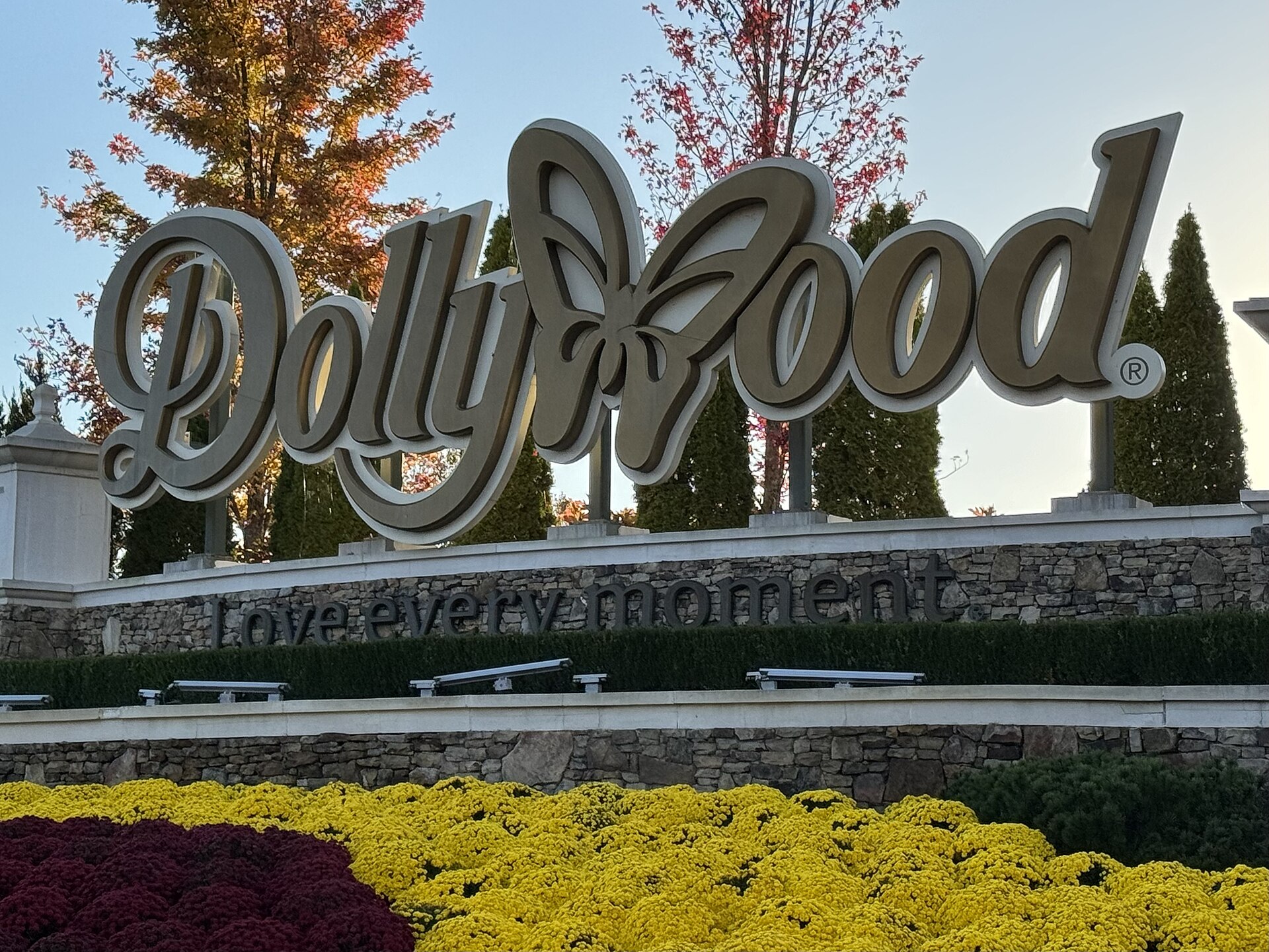The Dollywood entrance sign surrounded by red and orange fall trees and yellow and burgundy mums in Pigeon Forge, Tennessee